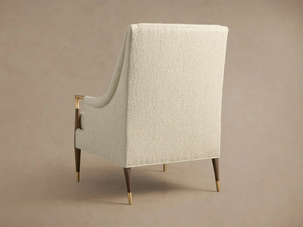 Close-up of the cream-colored Fisher High-Back Chair, featuring textured upholstery, brass armrests with dark wood, and a lumbar pillow.