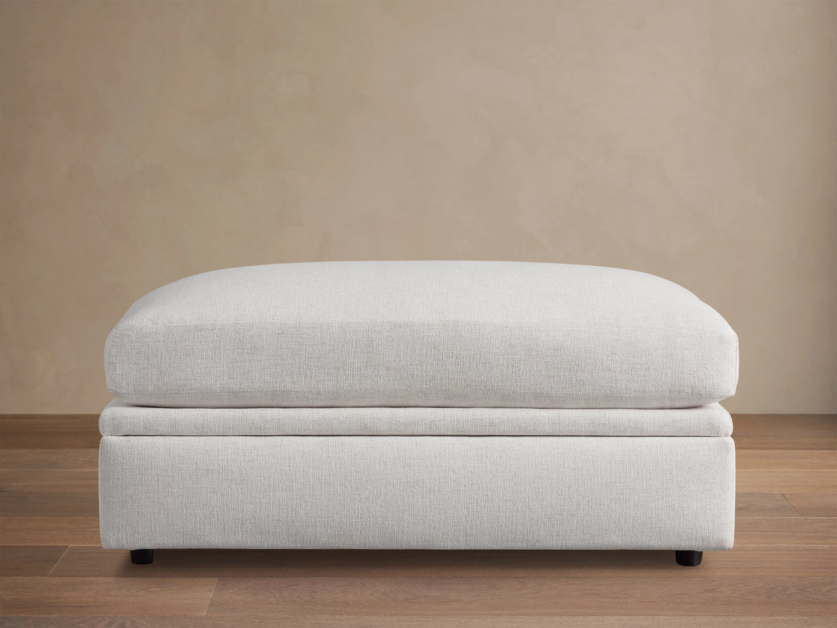 Light gray Kipton Storage Ottoman with smooth, cushioned top and base on dark wooden floor, set against a beige wall.