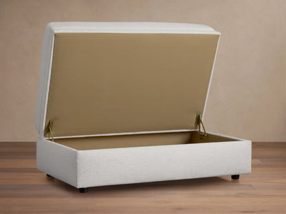 Light gray Kipton Storage Ottoman with smooth, cushioned top and base on dark wooden floor, set against a beige wall.
