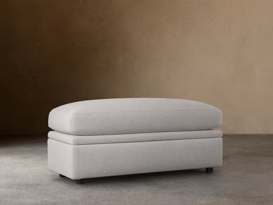 Light gray Kipton Storage Ottoman with smooth, cushioned top and base on dark wooden floor, set against a beige wall.