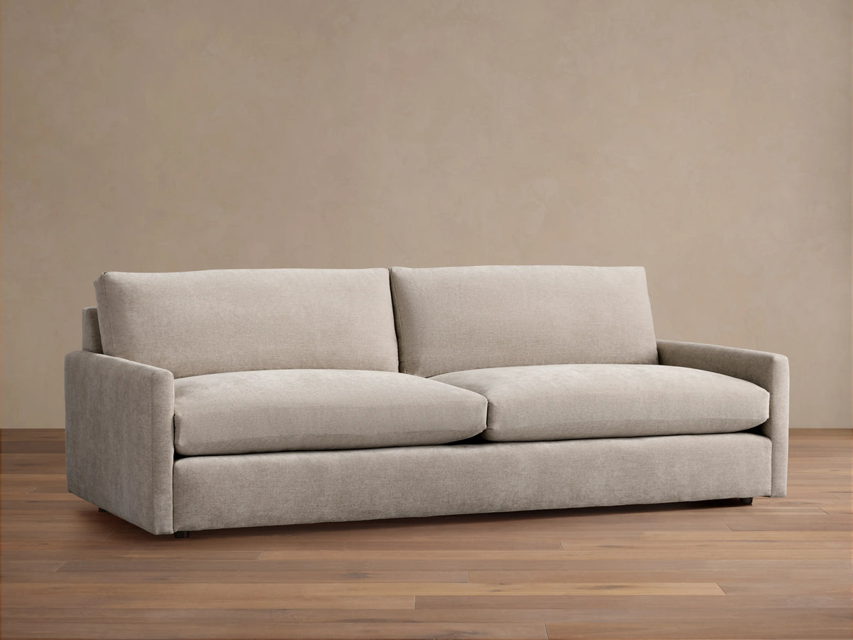 Kipton Sofa with a neutral beige fabric, featuring square arms and two large plush back cushions. The sofa is placed on a wooden floor.