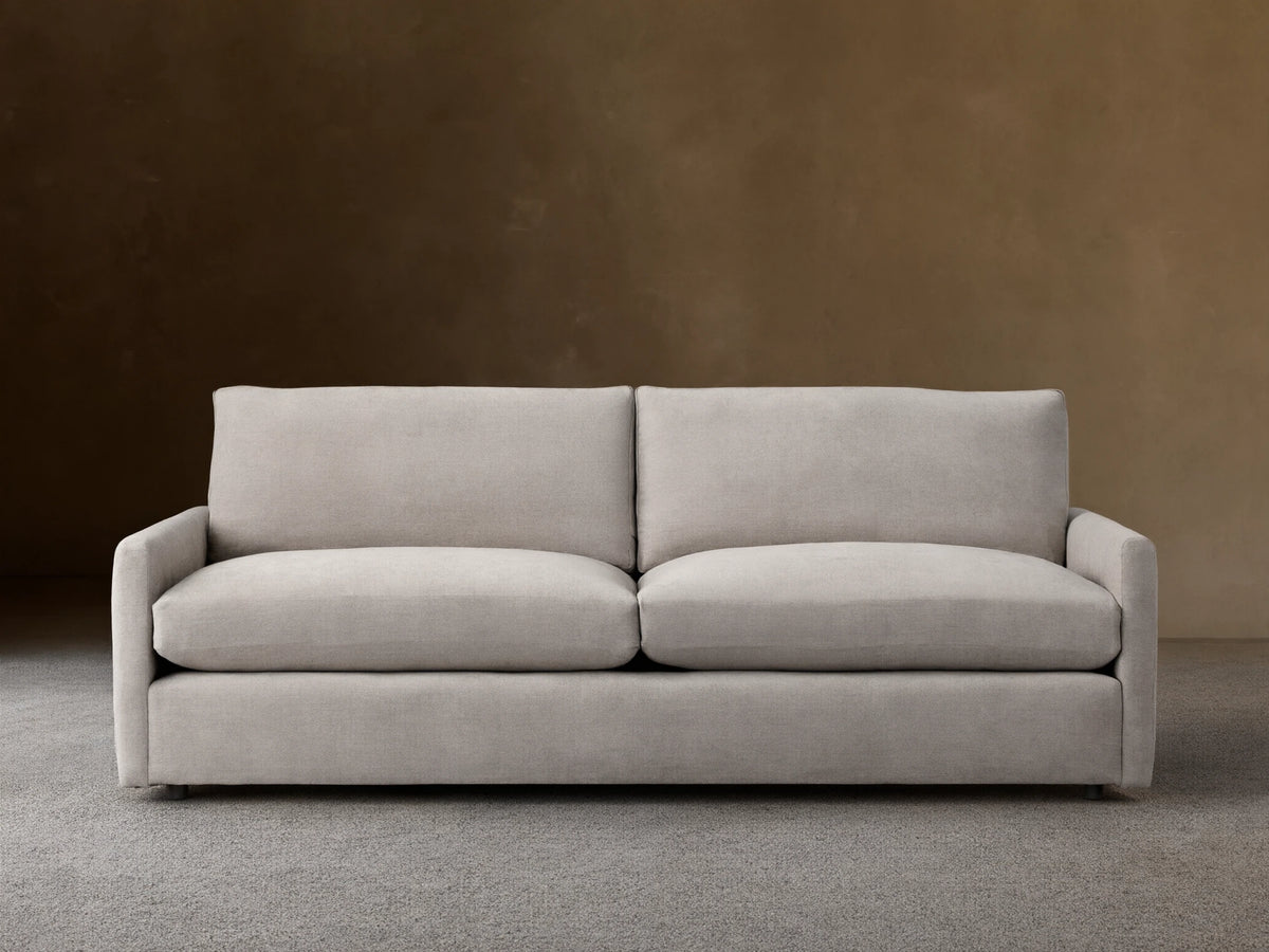 Kipton Sofa with a neutral beige fabric, featuring square arms and two large plush back cushions. The sofa is placed on a wooden floor.