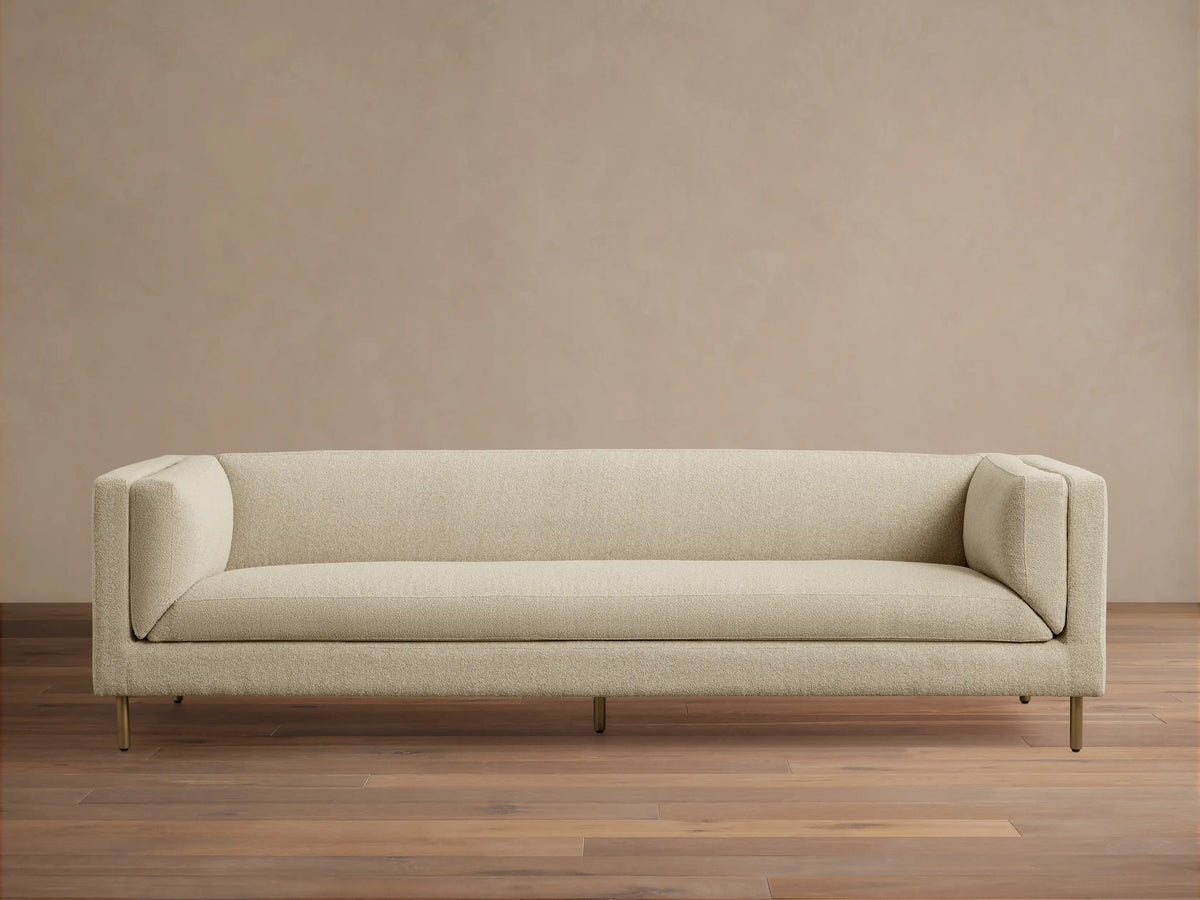 Modern beige textured fabric sofa with sleek lines, slender gold legs, against a tan wall on a wood floor.
