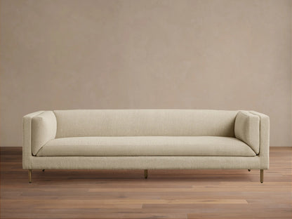 Modern beige textured fabric sofa with sleek lines, slender gold legs, against a tan wall on a wood floor.