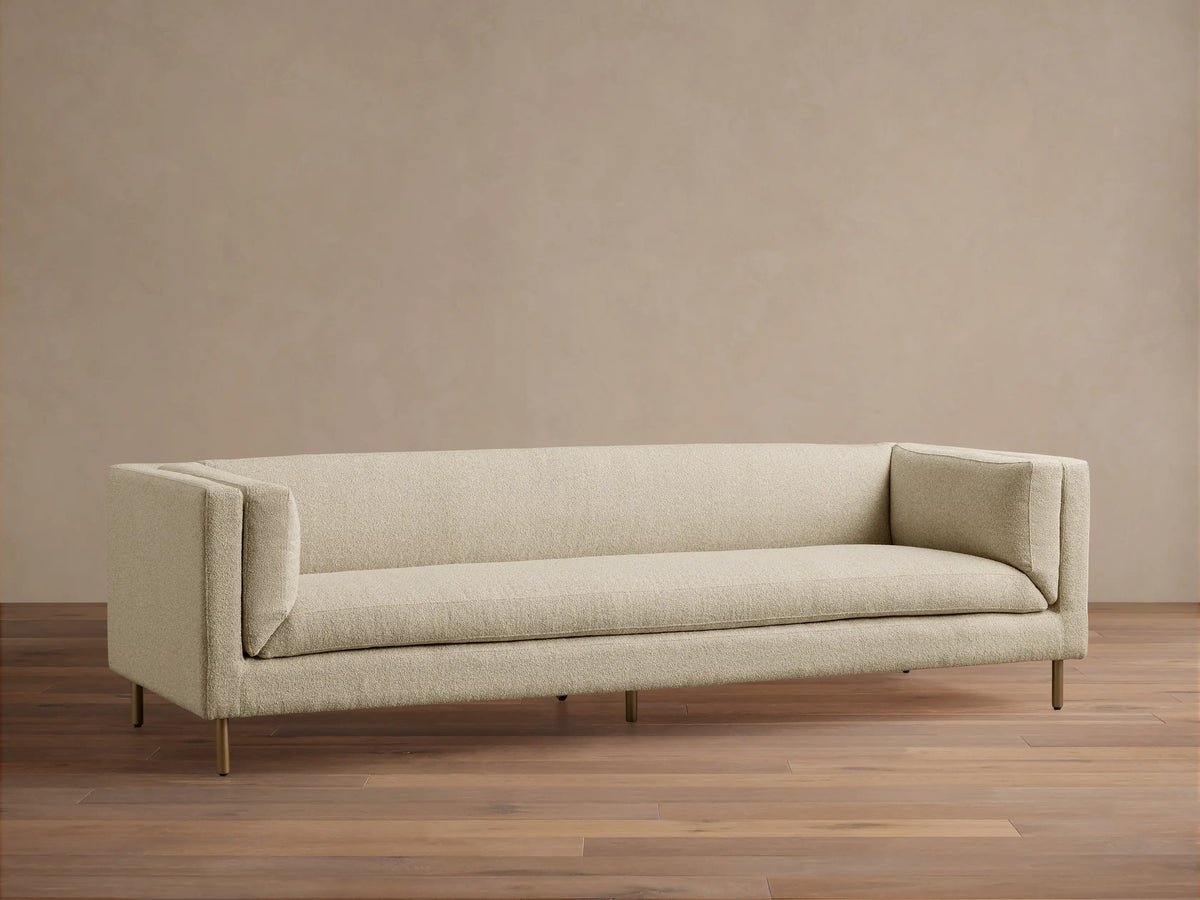 Modern beige textured fabric sofa with sleek lines, slender gold legs, against a tan wall on a wood floor.