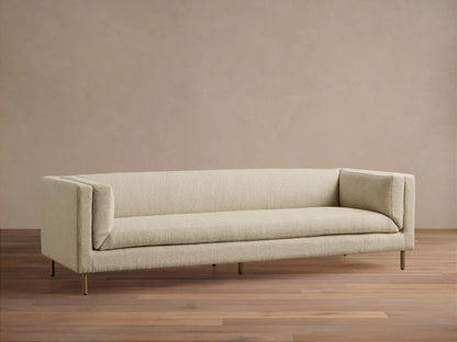 Modern beige textured fabric sofa with sleek lines, slender gold legs, against a tan wall on a wood floor.