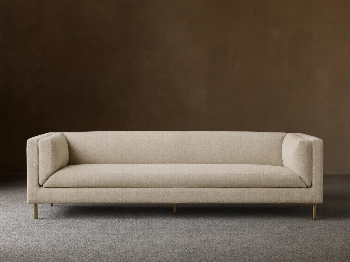 Modern beige textured fabric sofa with sleek lines, slender gold legs, against a tan wall on a wood floor.
