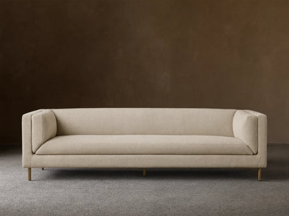 Modern beige textured fabric sofa with sleek lines, slender gold legs, against a tan wall on a wood floor.