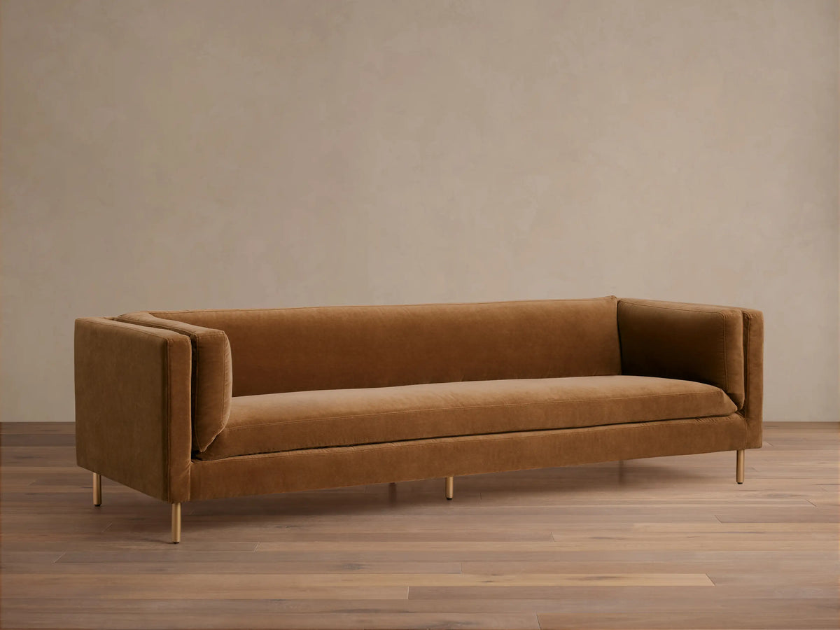 Warm brown velvet sofa with sleek square arms and golden legs, presented against a light wall and dark wood floor.