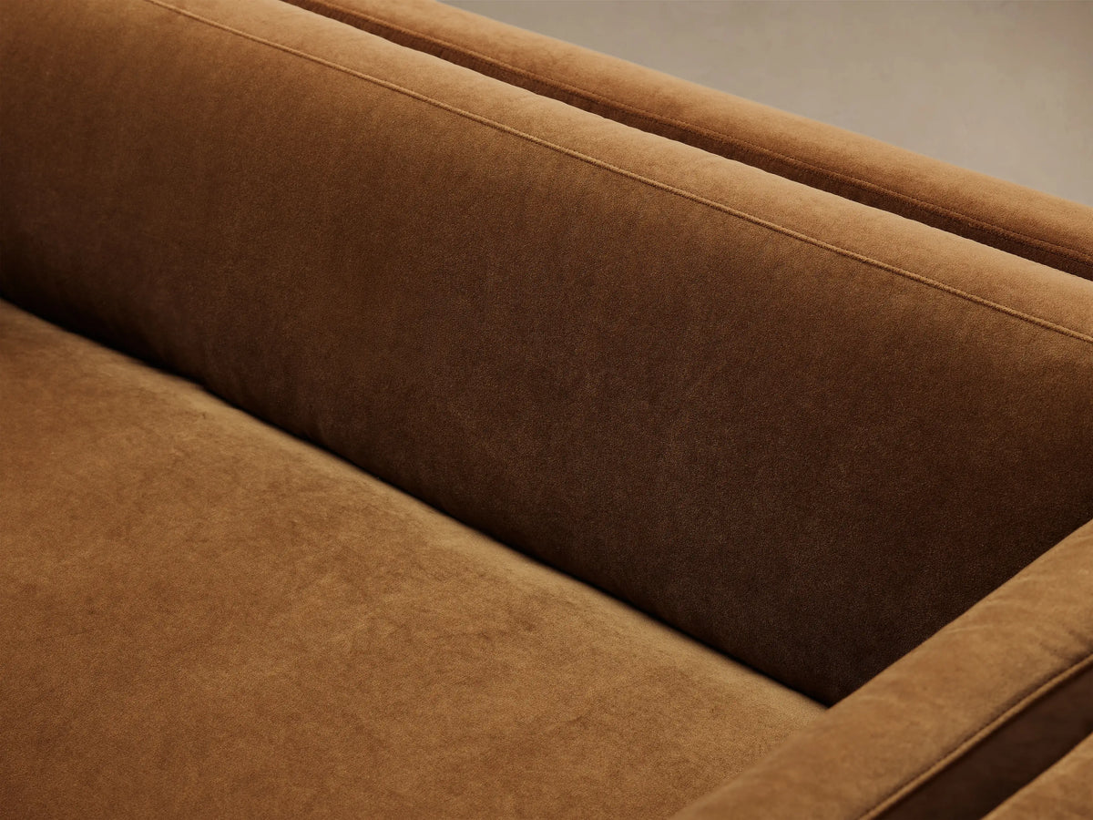 Warm brown velvet sofa with sleek straight arms, plump cushions, and slender gold legs, presented on a wooden floor.