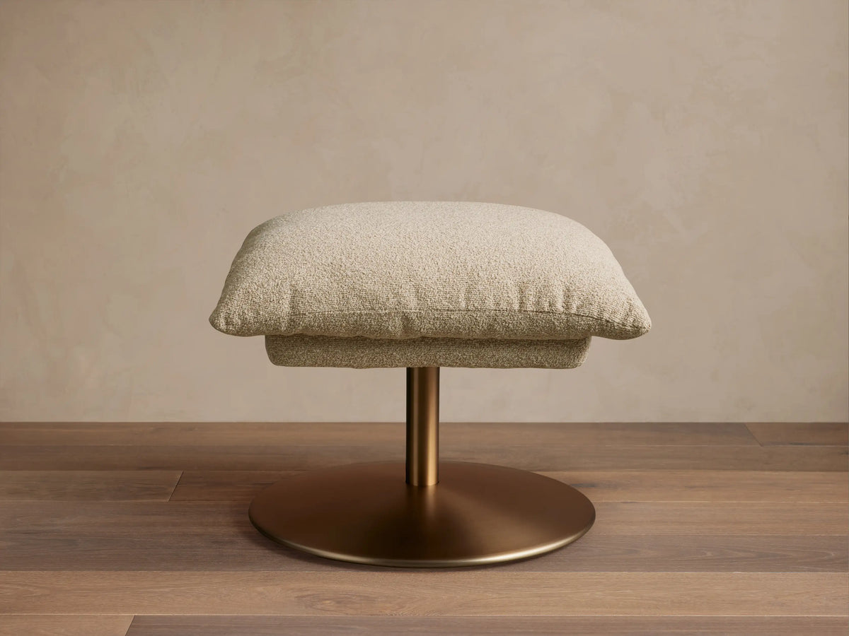 Beige Deacon Ottoman with cushioned top on a round bronze metal base, set on wooden floor, against a neutral wall.