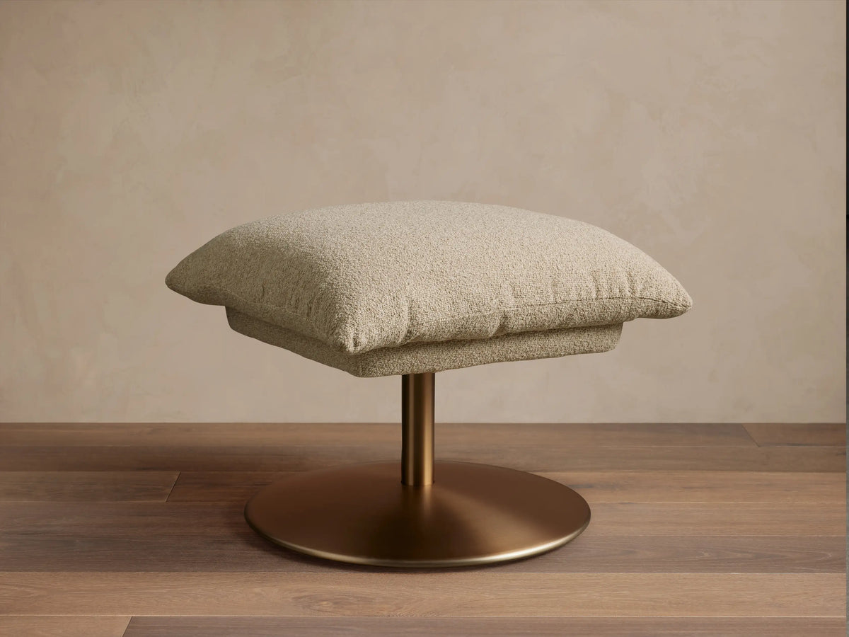 Beige Deacon Ottoman with cushioned top on a round bronze metal base, set on wooden floor, against a neutral wall.