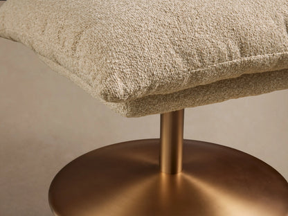 Deacon Ottoman with a textured beige cushion on a round, bronze-colored metal base, placed on a wooden floor.