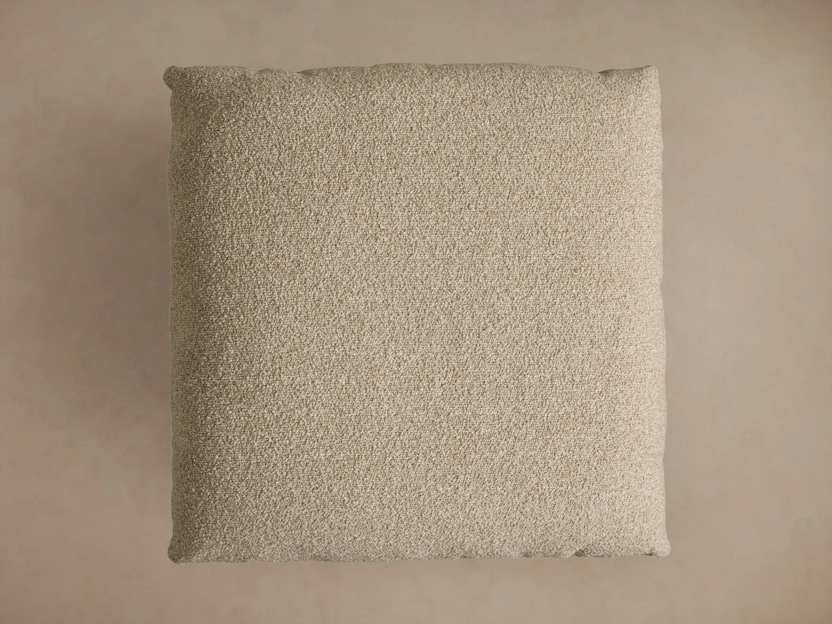 Deacon Ottoman with a textured beige cushion on a round, brushed bronze metal base, showcasing a modern design.
