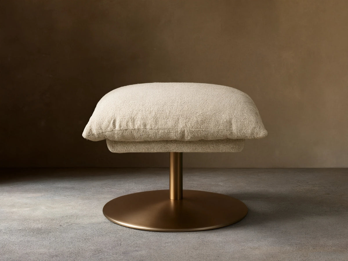 Beige Deacon Ottoman with cushioned top on a round bronze metal base, set on wooden floor, against a neutral wall.