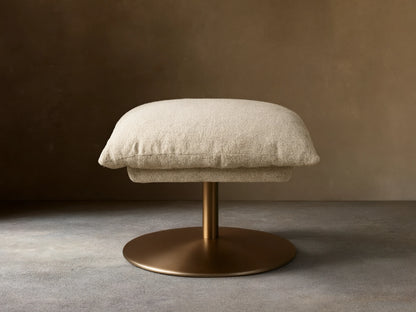 Beige Deacon Ottoman with cushioned top on a round bronze metal base, set on wooden floor, against a neutral wall.