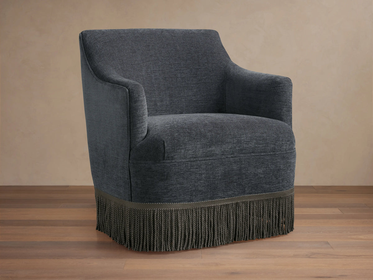 Savannah Swivel Chair in dark gray fabric, featuring a rounded back, sleek armrests, and a decorative fringe at the base.