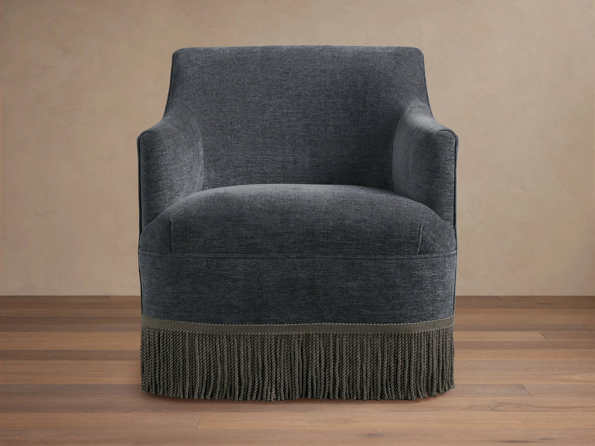 Savannah Swivel Chair in dark gray fabric, featuring a rounded back, sleek armrests, and a decorative fringe at the base.