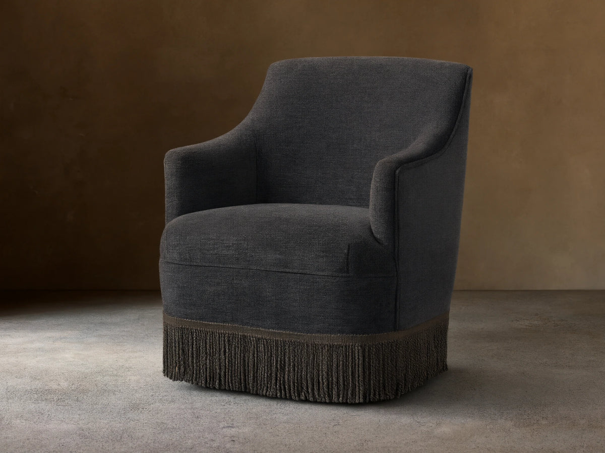 Savannah Swivel Chair in dark gray fabric, featuring a rounded back, sleek armrests, and a decorative fringe at the base.