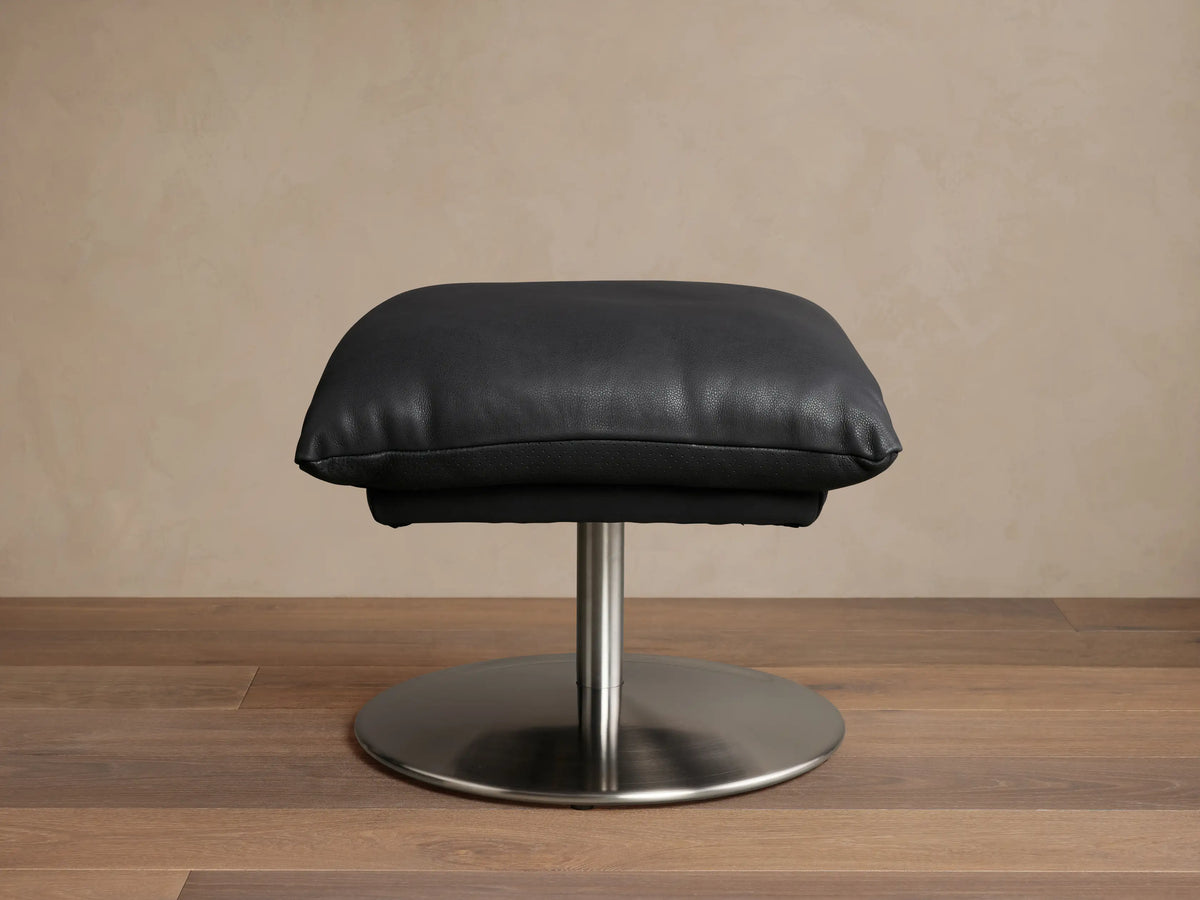 Deacon Leather Ottoman in black, featuring a plush cushion set on a sleek metal base, displayed on wooden flooring.