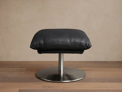 Deacon Leather Ottoman in black, featuring a plush cushion set on a sleek metal base, displayed on wooden flooring.