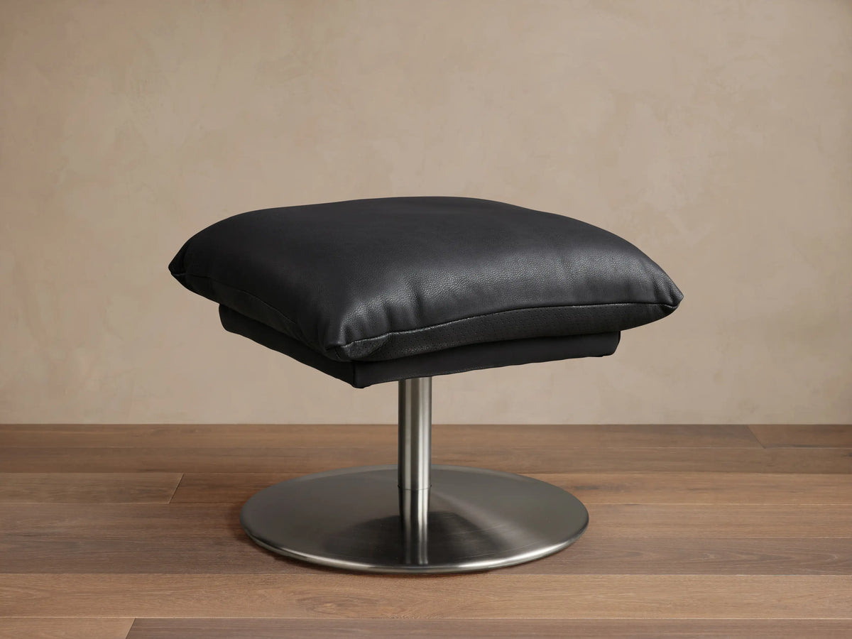 Deacon Leather Ottoman in black, featuring a plush cushion set on a sleek metal base, displayed on wooden flooring.