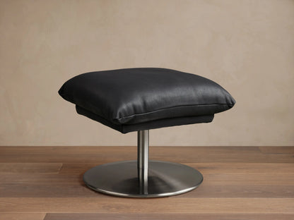 Deacon Leather Ottoman in black, featuring a plush cushion set on a sleek metal base, displayed on wooden flooring.