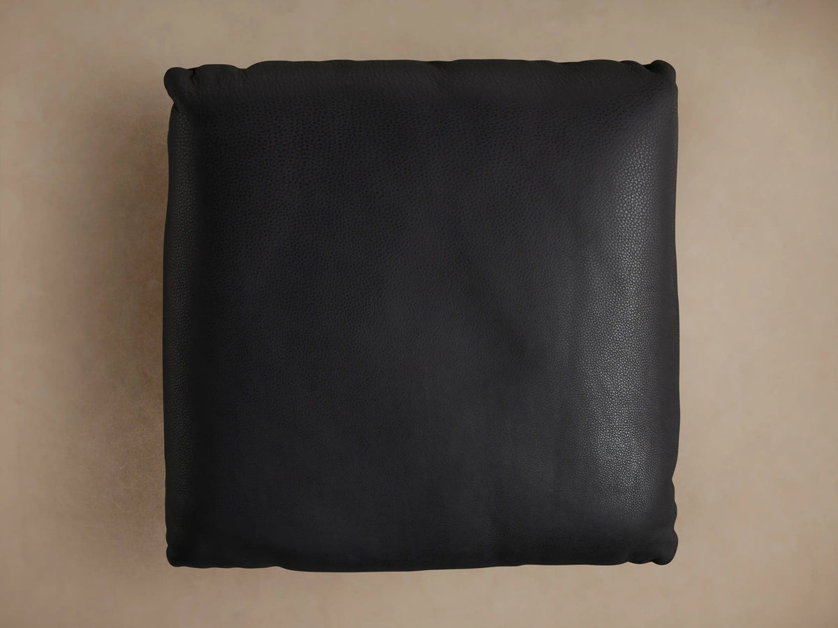 Deacon Leather Ottoman corner with black leather upholstery, detailed grain texture, and a round metal base.