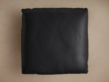 Deacon Leather Ottoman corner with black leather upholstery, detailed grain texture, and a round metal base.