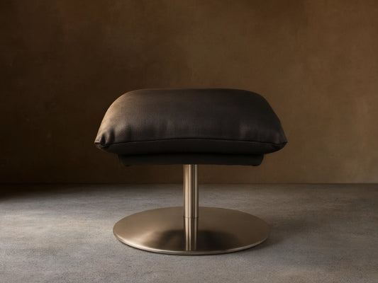 Deacon Leather Ottoman in black, featuring a plush cushion set on a sleek metal base, displayed on wooden flooring.
