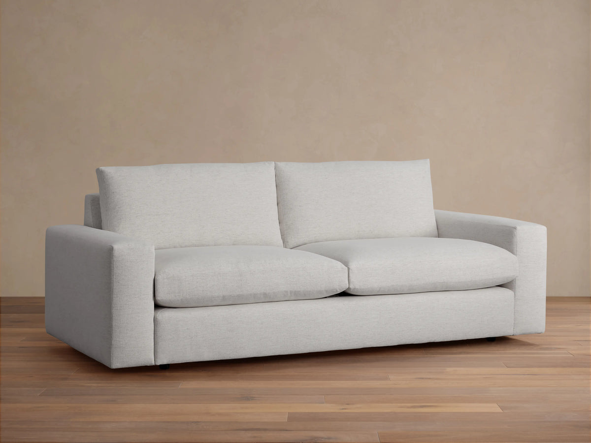 Kipton Wide Arm Sofa in light gray fabric, featuring wide arms and deep cushioning, on a wooden floor with a neutral background.