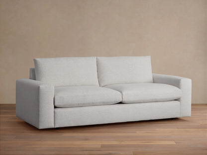 Kipton Wide Arm Sofa in light gray fabric, featuring wide arms and deep cushioning, on a wooden floor with a neutral background.