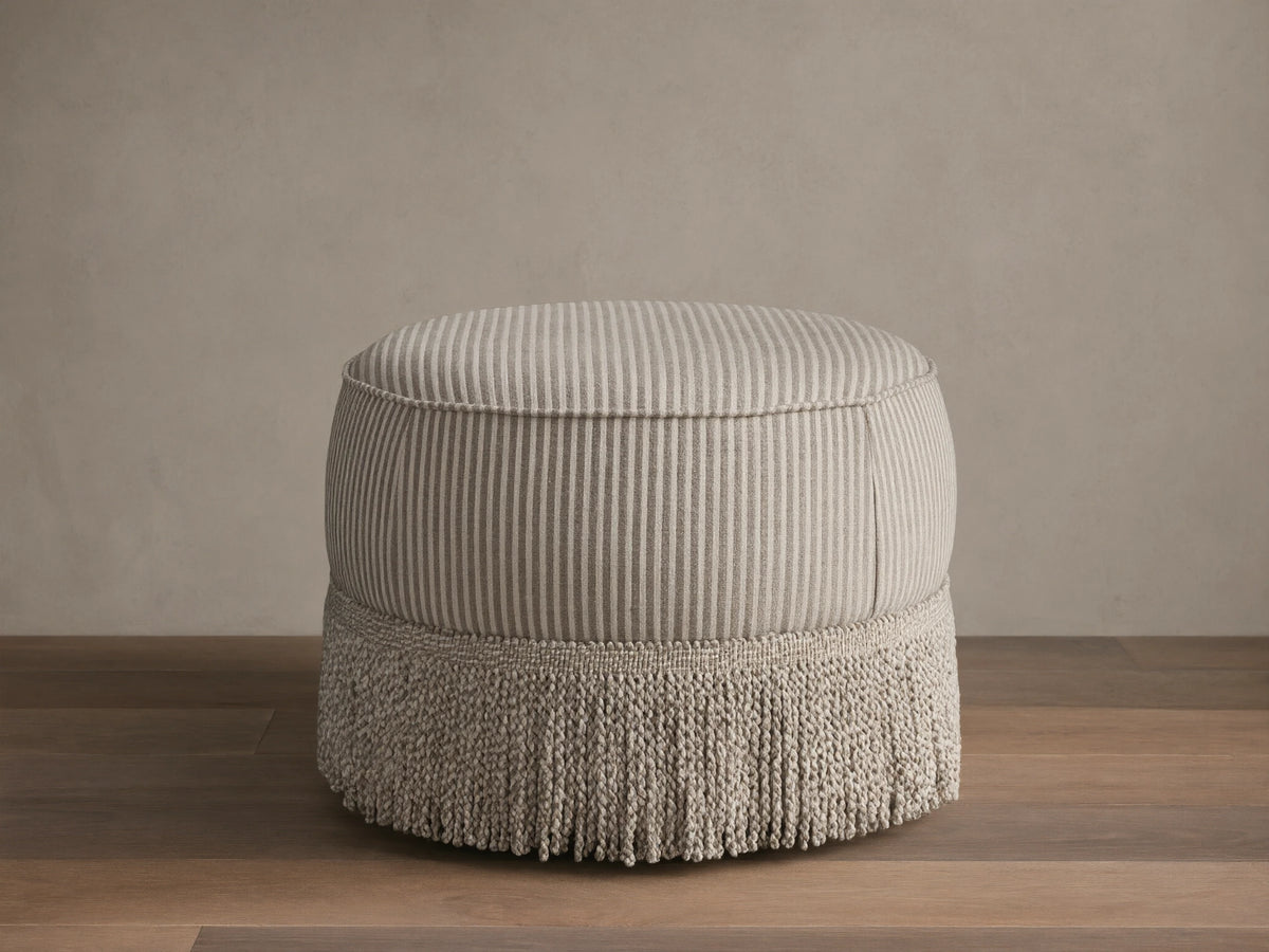Front view of the round Mimi Ottoman, upholstered in beige and taupe striped fabric with a matching fringe base.