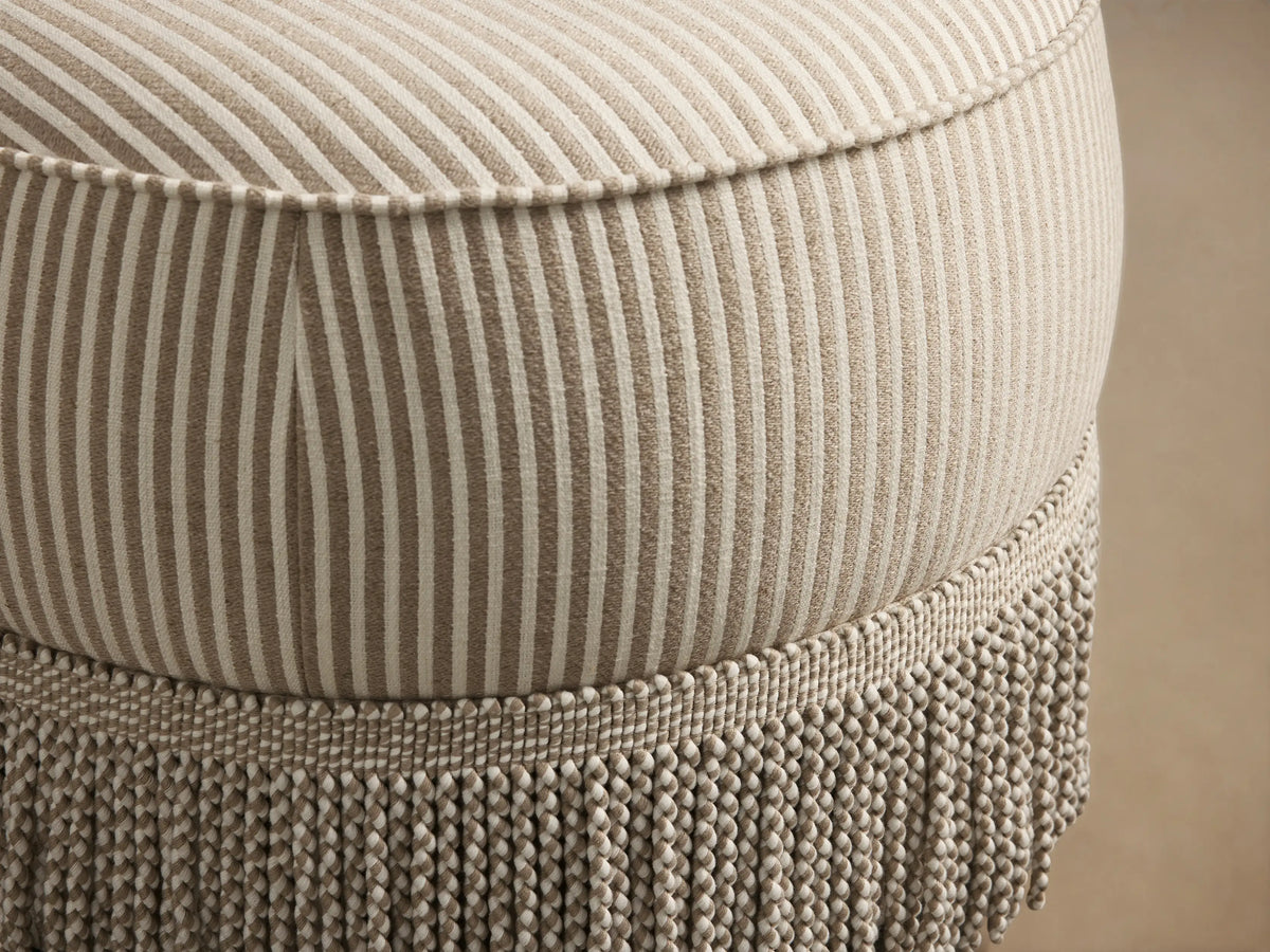 Close-up of the beige and off-white pinstriped Mimi Ottoman, showing its rounded top and textured fringe trim.