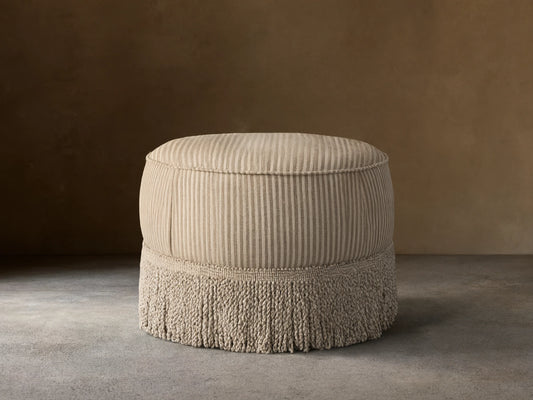 Front view of the round Mimi Ottoman, upholstered in beige and taupe striped fabric with a matching fringe base.