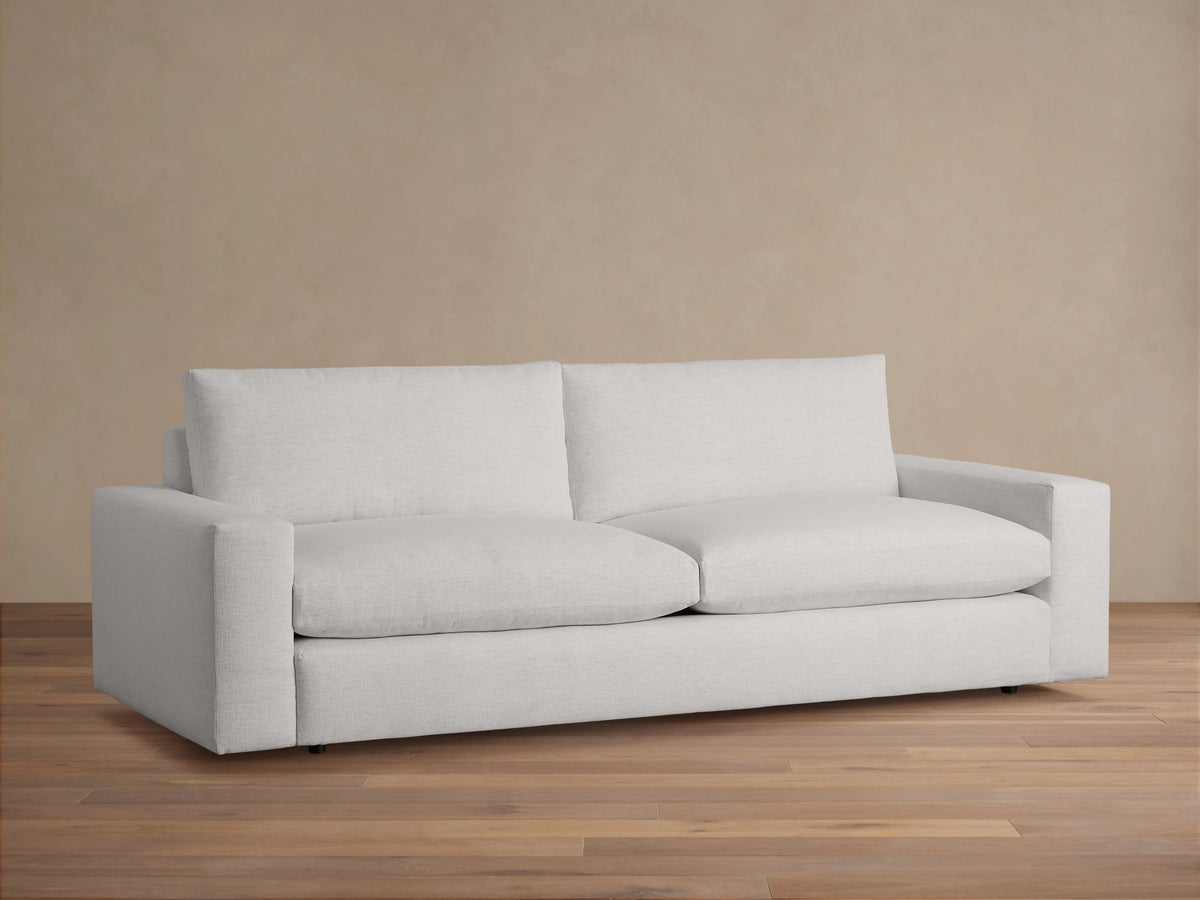 Wide-arm sofa with light gray upholstery, featuring thick seat and back cushions, placed on a wooden floor against a beige wall.