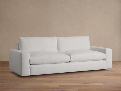 Wide-arm sofa with light gray upholstery, featuring thick seat and back cushions, placed on a wooden floor against a beige wall.