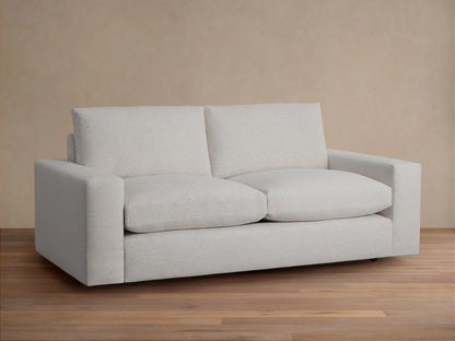 Kipton Wide Arm Sofa in light beige fabric, featuring two large seat cushions and two backrests. Set on a wooden floor against a neutral wall.