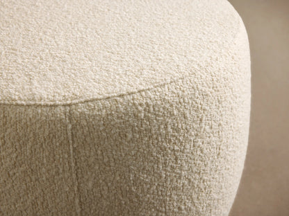 Ryles Ottoman: Round, cream boucle ottoman sitting on a dark wood floor against a light tan wall.