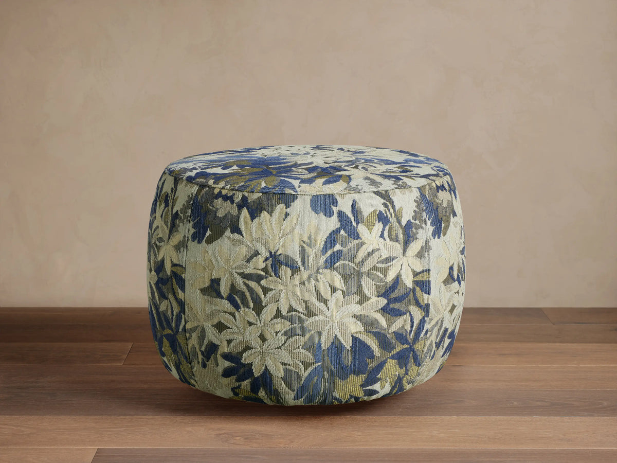 Ryles Ottoman, round, upholstered in blue, green, and cream floral fabric, on a wooden floor against a beige wall.