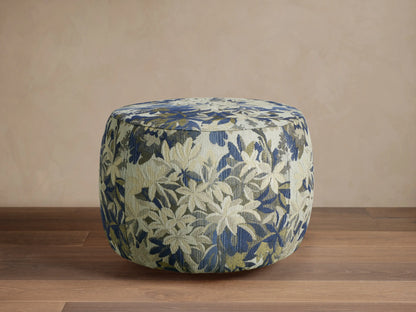 Ryles Ottoman, round, upholstered in blue, green, and cream floral fabric, on a wooden floor against a beige wall.