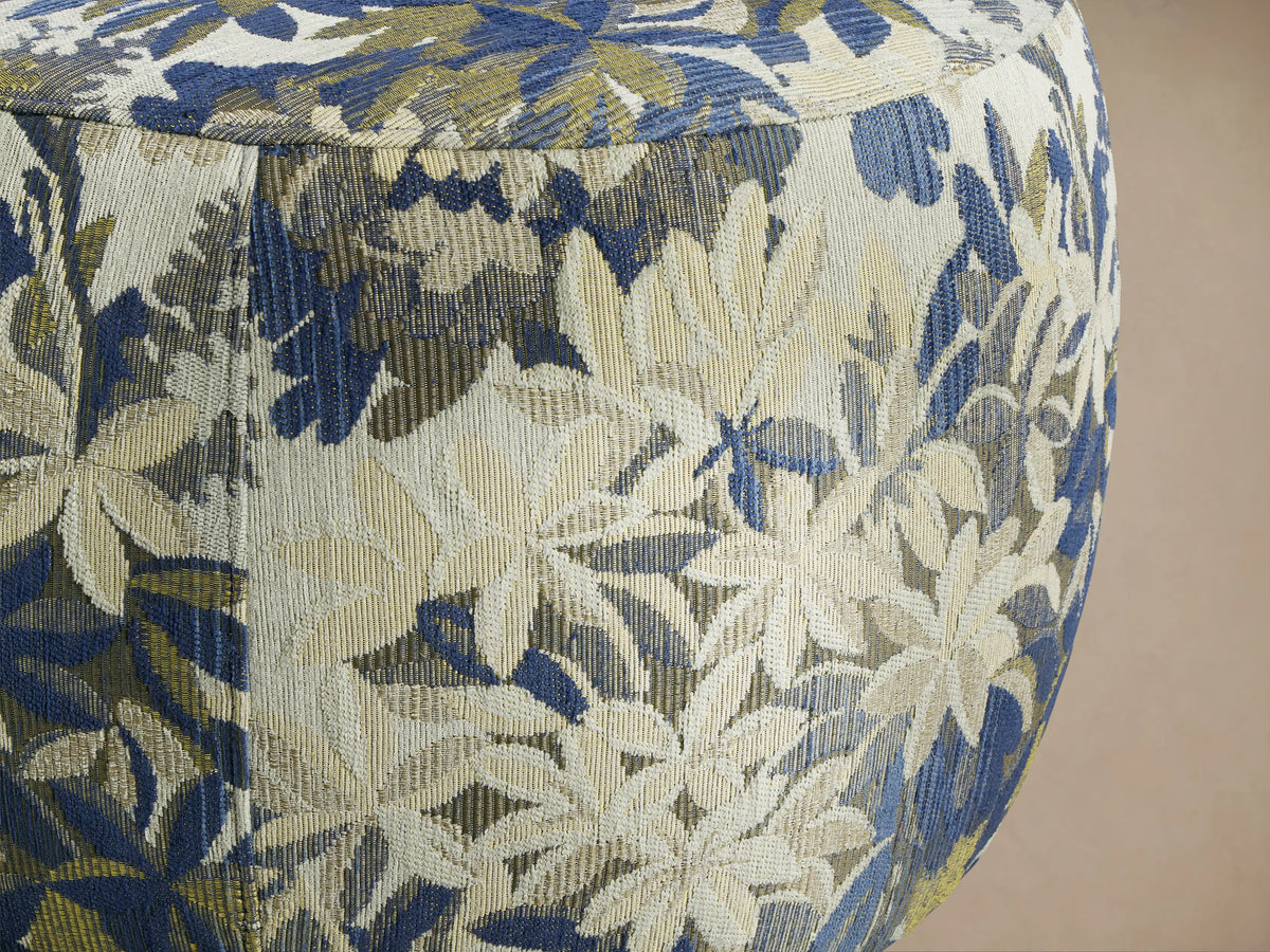 Ryles Ottoman, round, upholstered in blue, green, and cream floral fabric, on a wooden floor against a beige wall.
