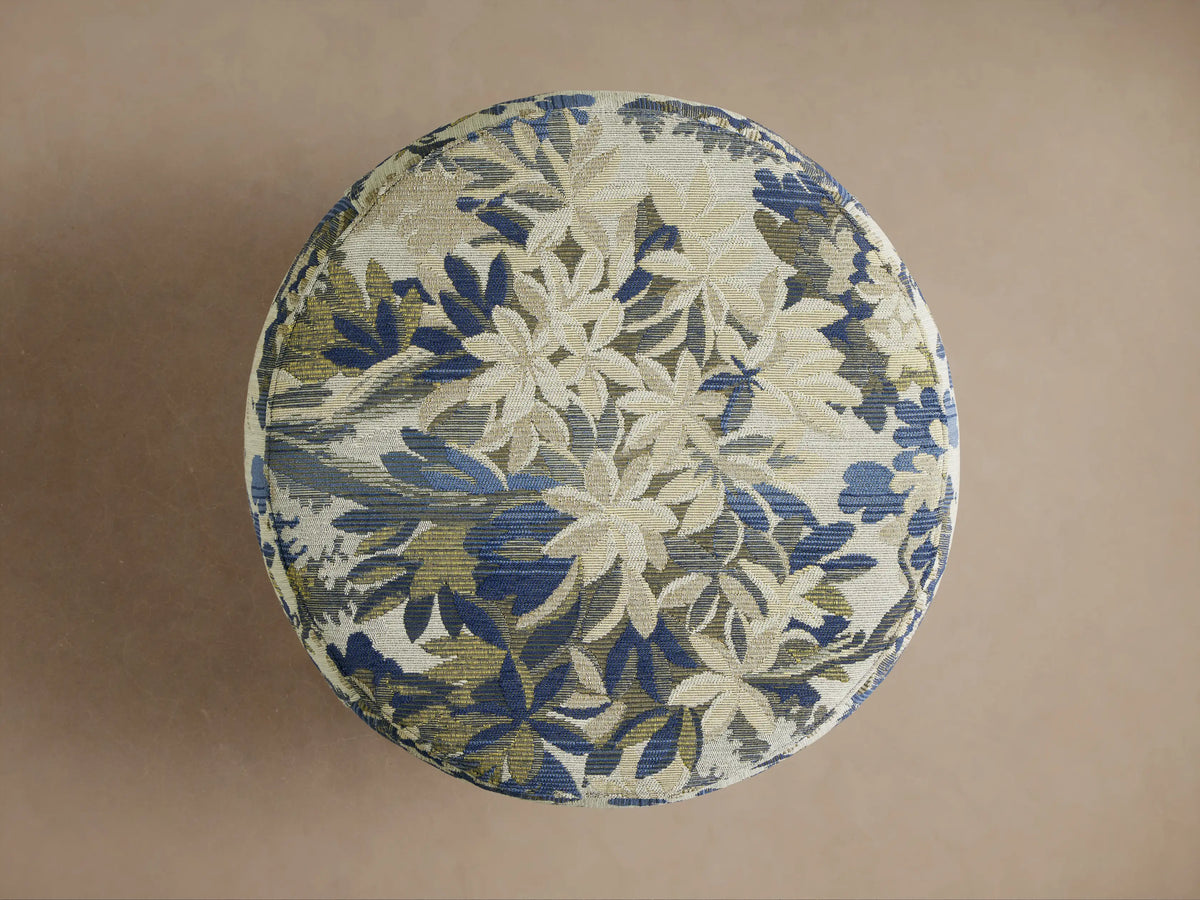 Close-up of a round Ryles Ottoman upholstered in textured fabric with a floral pattern in shades of blue, gold, and olive green.