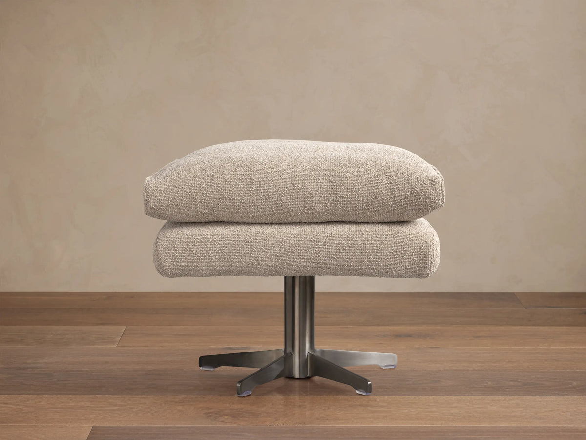 Giorgio Ottoman: Two stacked, light beige Abi Fawn fabric cushions on a brushed metal four-star base, on a wooden floor.
