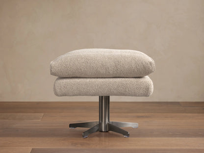 Giorgio Ottoman: Two stacked, light beige Abi Fawn fabric cushions on a brushed metal four-star base, on a wooden floor.