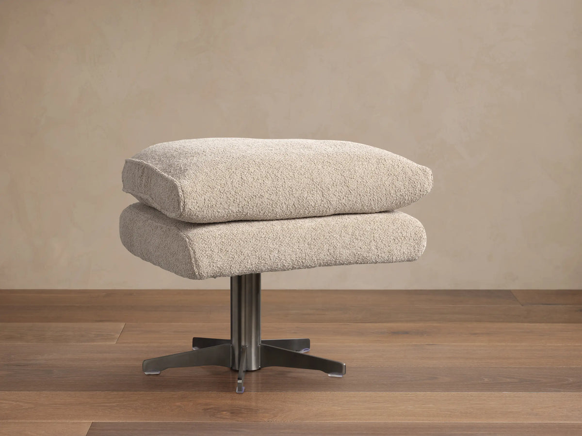 Giorgio Ottoman: Two stacked, light beige Abi Fawn fabric cushions on a brushed metal four-star base, on a wooden floor.