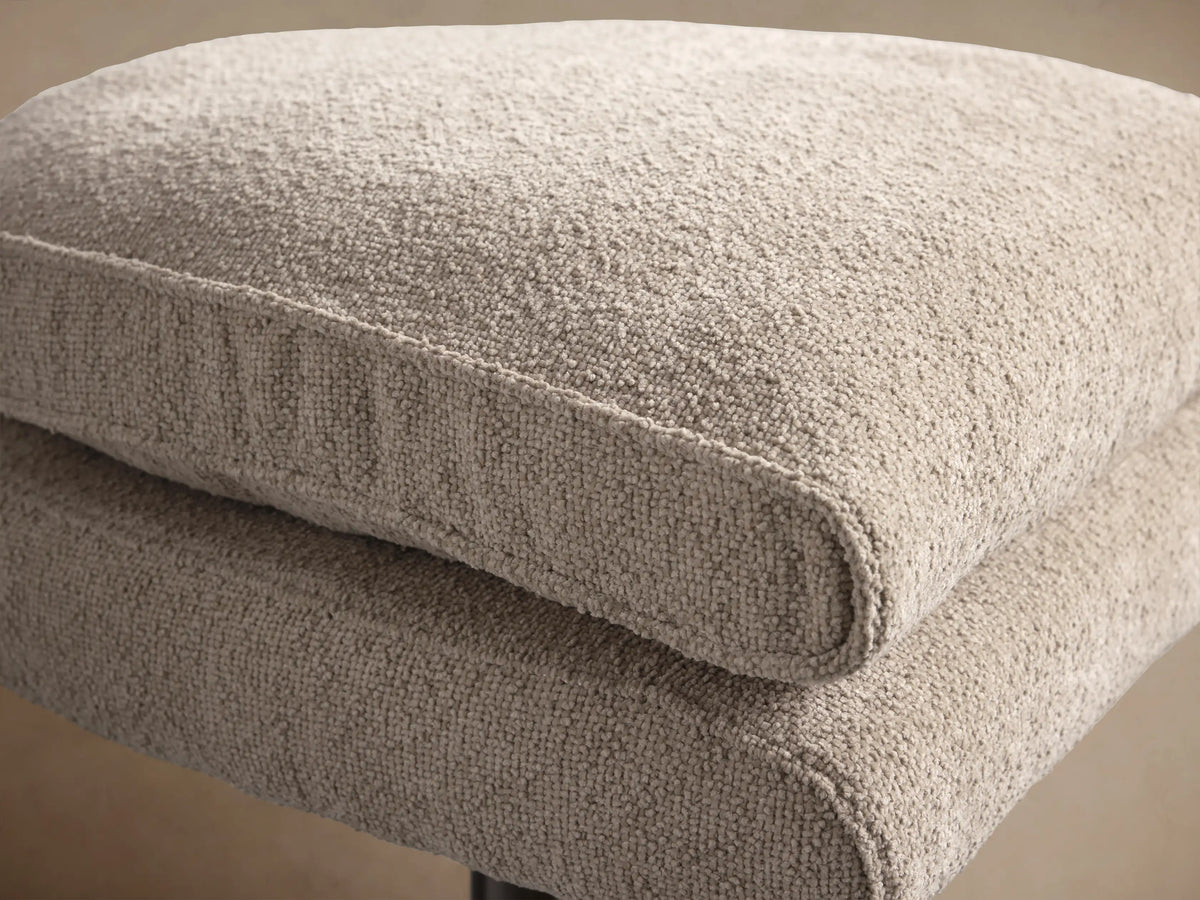 Giorgio Ottoman: Two stacked rectangular cushions in light fawn textured fabric on a brushed metal star base, on a wooden floor.