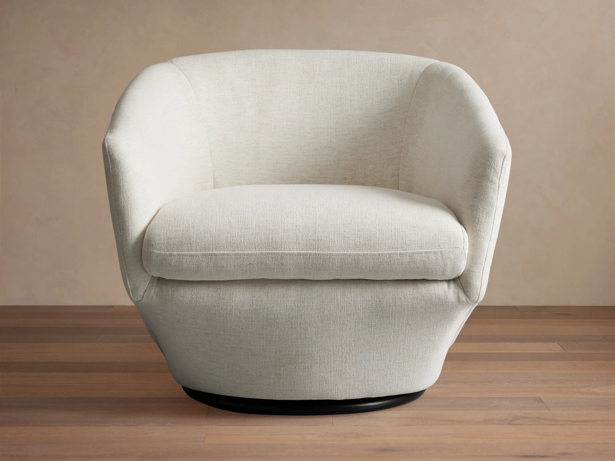 Swivel chair with light beige fabric, curved armrests, cushioned seat and backrest on a black circular base, sitting on wood floor.