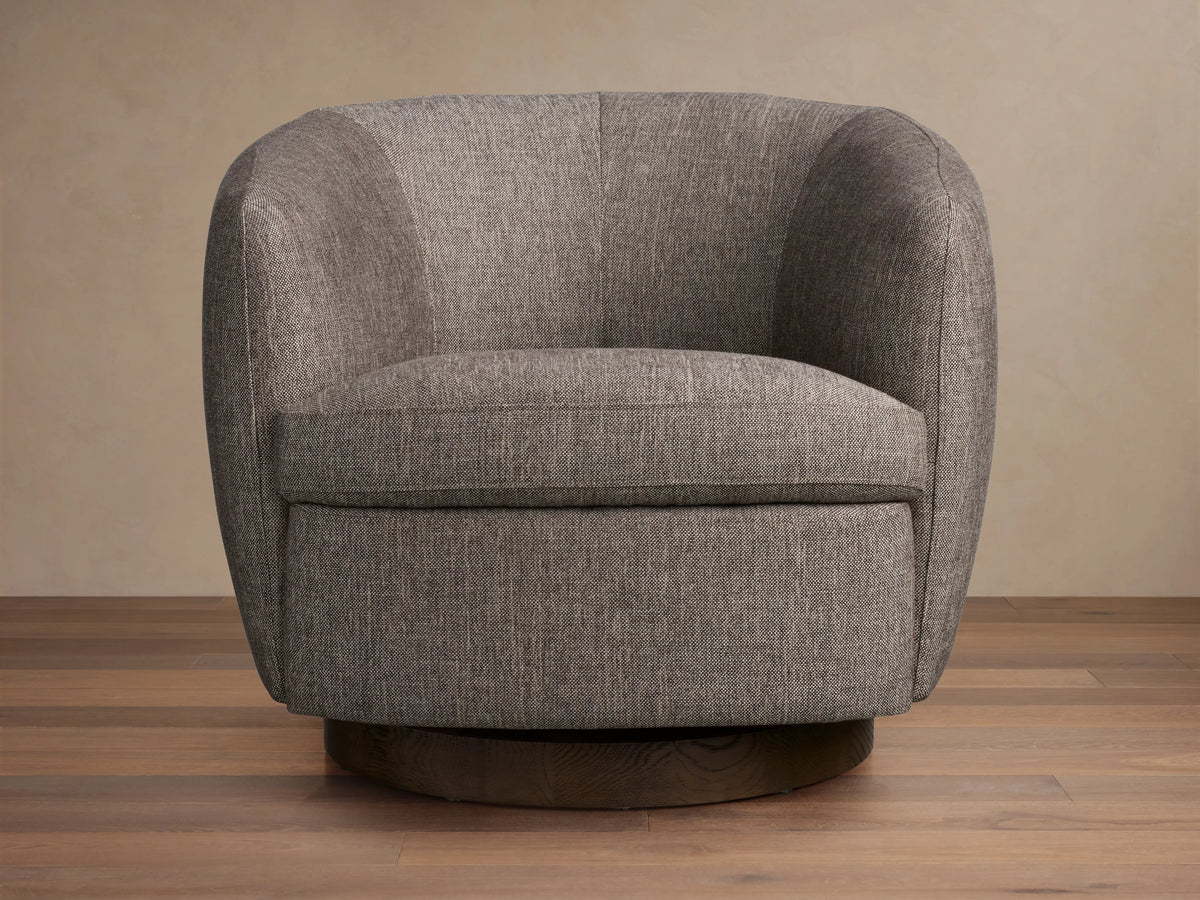 Cerrito Swivel Chair in a soft grey fabric, featuring a rounded backrest, plush cushioning, and a wooden swivel base, on a wooden floor.