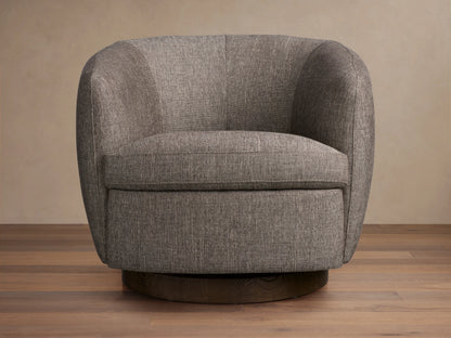 Cerrito Swivel Chair in a soft grey fabric, featuring a rounded backrest, plush cushioning, and a wooden swivel base, on a wooden floor.